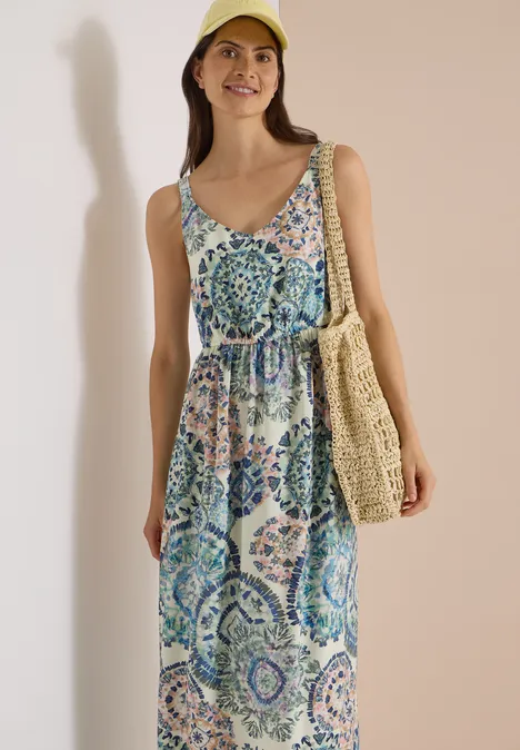 Eine lächelnde Frau trägt ein bunt gemustertes, ärmelloses Maxikleid, eine Strohtasche und eine gelbe Mütze.