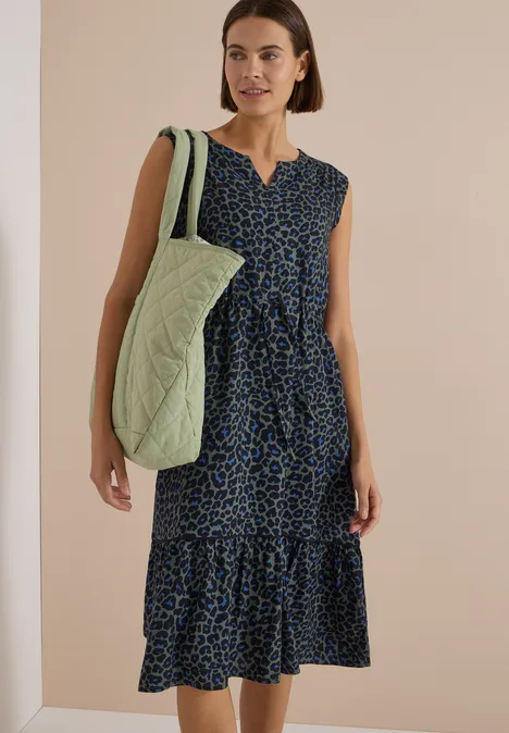 Frau trägt midi-Kleid ohne Ärmel mit blau-schwarzem Muster, V-Ausschnitt und grüner, gesteppter Schultertasche.