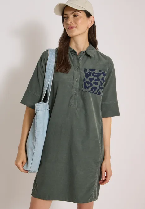 Frau in olivgrünem Polokleid mit Leopardenmuster-Tasche, hellblauer Stofftasche über der Schulter und Basecap.