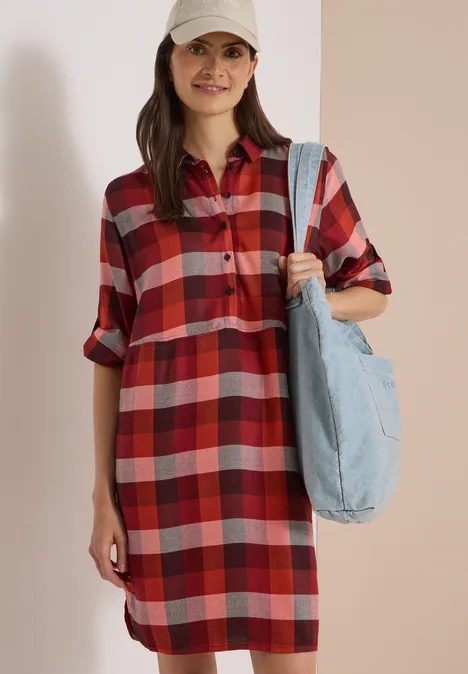 Frau in rotem kariertem Hemdkleid mit aufgekrempelten Ärmeln, heller Baseballmütze und Jeans-Tasche über der Schulter.