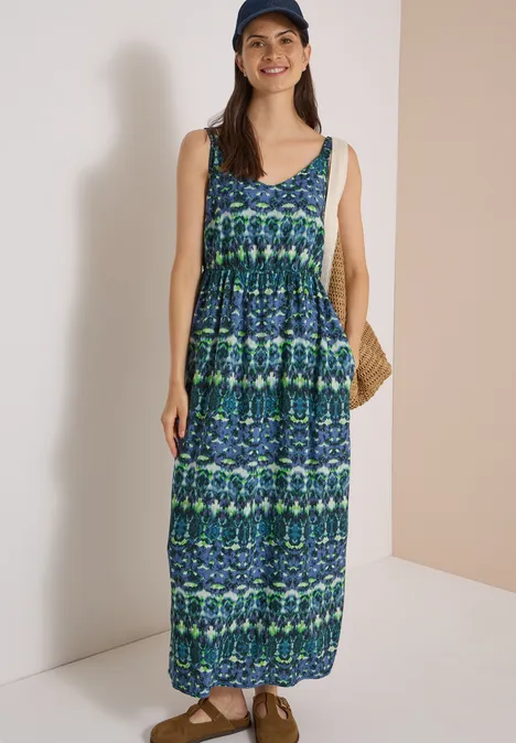 Maxikleid mit V-Neck und Print urban dark blue Frau in blau-grünem Maxikleid, mit Strohtasche und Baseballcap, steht lächelnd vor neutralem Hintergrund.