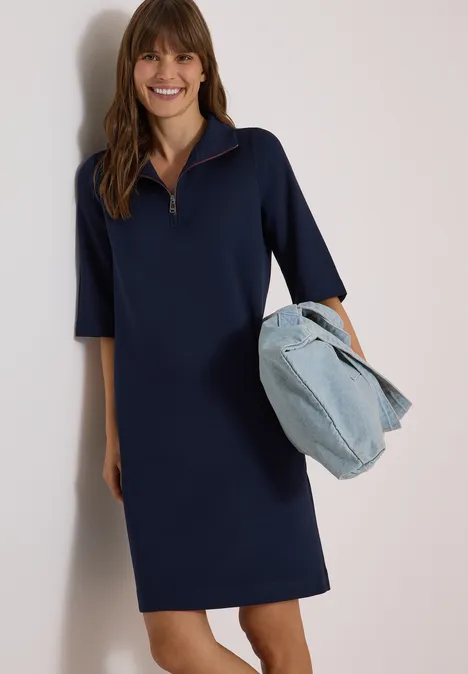 Frau trägt ein navyblaues Kleid und hält eine helle Jeans-Tasche, sie lächelt.