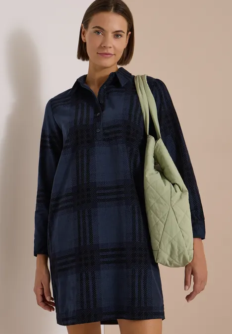 Frau trägt dunkelblau-schwarz kariertes Hemdkleid und eine hellgrüne, gesteppte Schultertasche.
