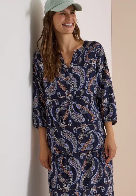 Frau in blauem Paisley-Oberteil und passendem Rock lächelt.