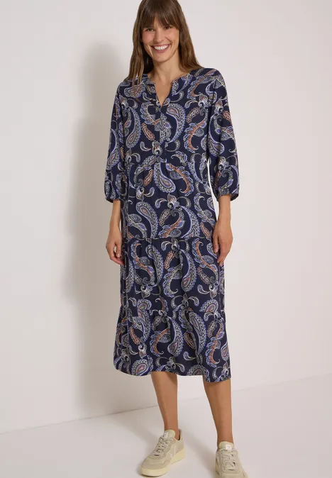 Frau trägt midi-langes blaues Paisley-Kleid mit V-Ausschnitt, 3/4-Ärmeln und hellen Sneakern.