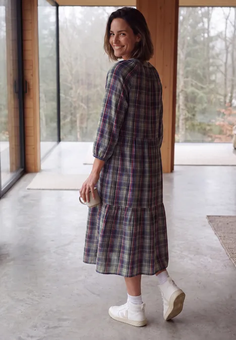Frau im karierten Kleid, weiße Socken und Sneaker, hält Tasse und lächelt am Fenster.