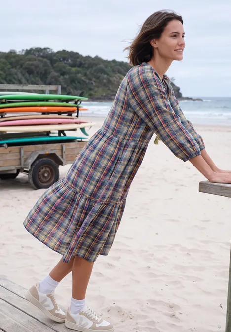 Une femme appuyée sur une rambarde de plage, vêtue d’une robe à carreaux, chaussettes et baskets, surfeurs en arrière-plan.