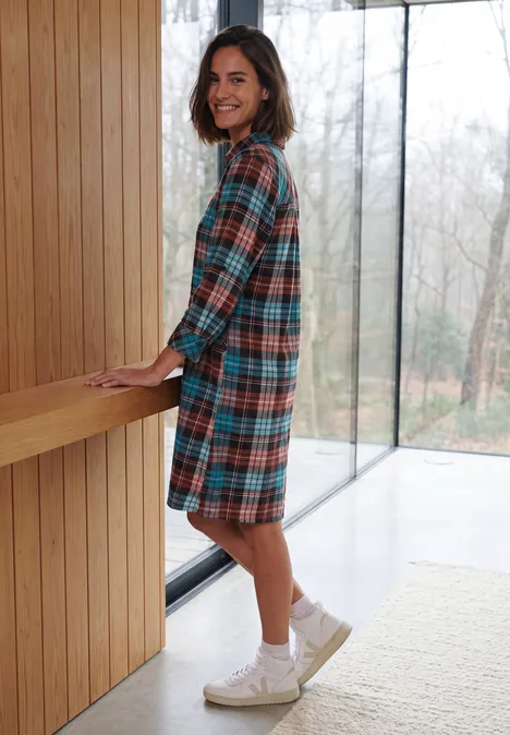 Eine lächelnde Frau in einem karierten Hemdblusenkleid lehnt an einer Holztheke vor großen Glastüren.
