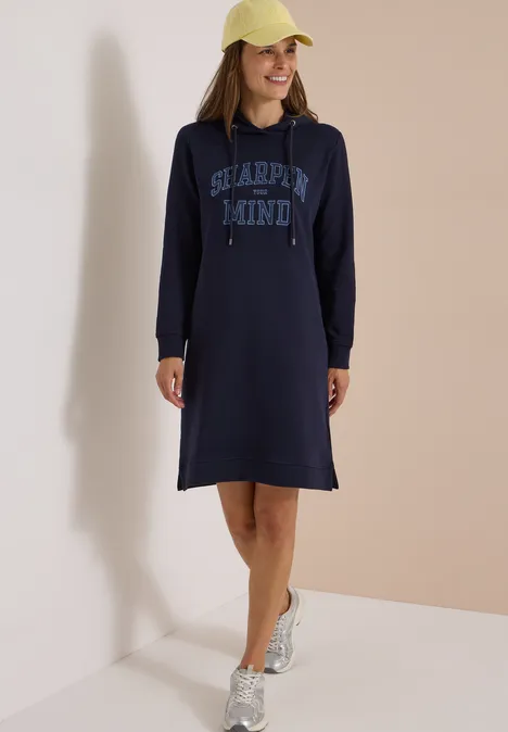 Eine lächelnde Frau trägt ein dunkelblaues Hoodie-Kleid mit Kapuze und Aufschrift, kombiniert mit Kappe und Sneakern.