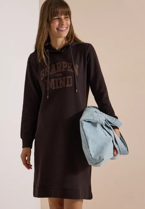 Frau lächelt, trägt braunes Hoodie-Kleid mit Aufdruck und hält eine hellblaue Tasche.