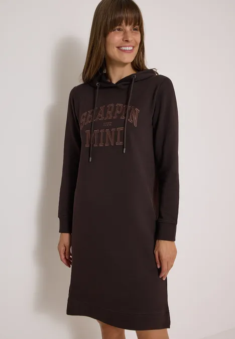 Frau trägt ein braunes Hoodie-Kleid mit Kapuze und Print, lächelt.