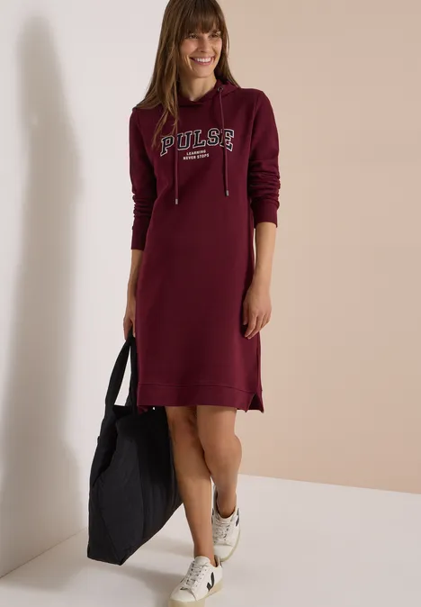Frau in burgunderfarbenem Kapuzenkleid mit "Poler"-Schriftzug, hält schwarze Tasche und trägt weiße Sneaker.