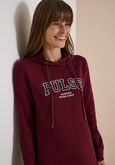 Frau mit langem braunem Haar lächelt und trägt burgunderfarbenen Hoodie mit Aufschrift "PULSE".