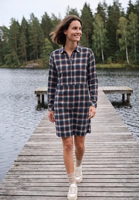 Knielanges Kleid mit Karomuster urban dark blue Eine Frau in kariertem Hemdblusenkleid spaziert lächelnd auf einem Holzsteg am ruhigen See, umgeben von Wald.
