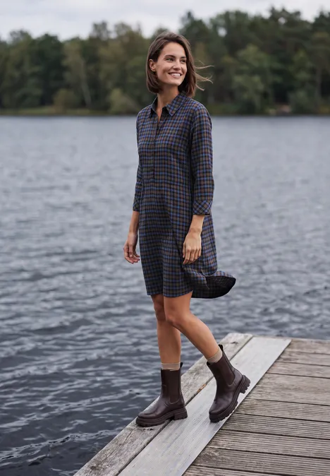 Frau in kariertem Hemdblusenkleid und Gummistiefeln spaziert lächelnd auf einem Steg am See.