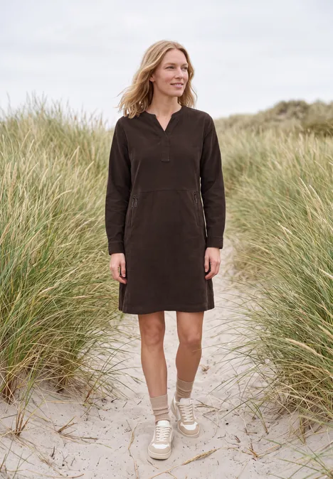 Knielanges Kleid aus Cord mit Split Neck truffle brown Frau in einem langen braunen Kleid steht an einem sandigen Strand mit Dünen