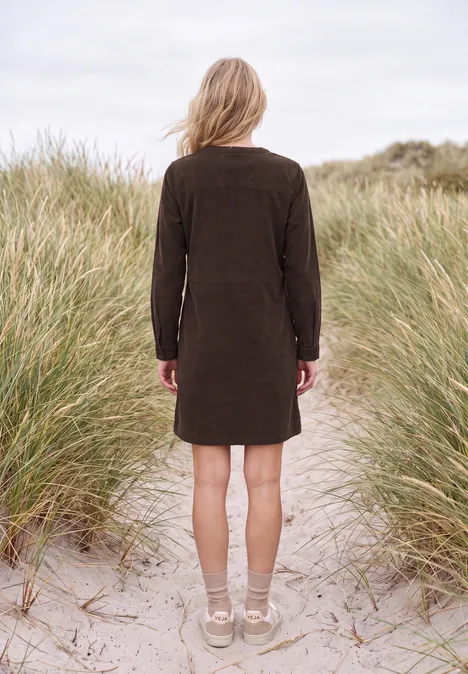 Knielanges Kleid aus Cord mit Split Neck truffle brown Frau in braunem Kleid steht mit dem Rücken zum Betrachter auf sandigen Dünen zwischen Strandgras.