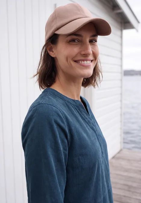 Lächelnde Frau mit Baseballcap und blauem Oberteil steht am Wasser vor einem weißen Gebäude.