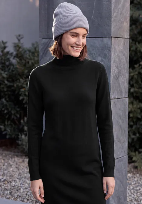 Eine lächelnde Frau in schwarzem Rollkragenkleid und grauer Mütze steht draußen neben einem Steinpfeiler und Pflanzen.