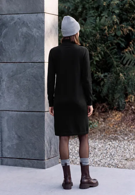 Eine Frau im schwarzen Strickkleid, Stiefeln und Mütze steht mit dem Rücken zur Kamera vor Naturkulisse