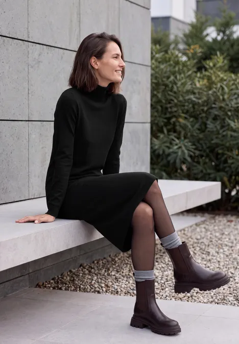 Frau in schwarzem Rollkragenkleid sitzt auf Bank im Freien, lächelt, trägt graue Socken und braune Stiefel.