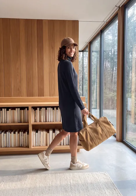 Robe-pull longueur genou à col roulé urban dark blue Femme en robe bleue et casquette portant un sac dans un intérieur moderne avec bois et baies vitrées.