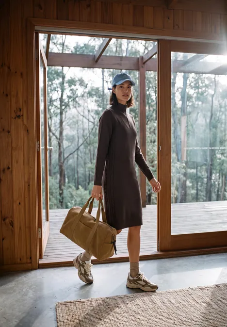 Femme en robe tricotée et baskets, sac de voyage, devant de larges fenêtres donnant sur la forêt.