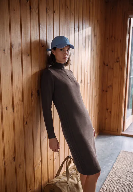 Femme appuyée contre un mur en bois, portant une robe marron et une casquette bleue, à côté un sac.
