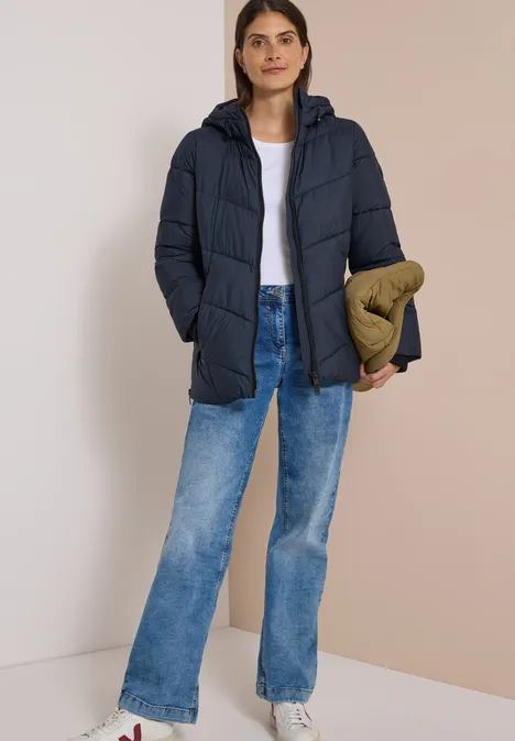 Puffer-Jacke in Midi-Länge mit Kapuze dark navy blue Frau in dunkelblauer Steppjacke, weißem Shirt, blauen Jeans, hält eine olivgrüne Tasche vor neutralem Hintergrund.