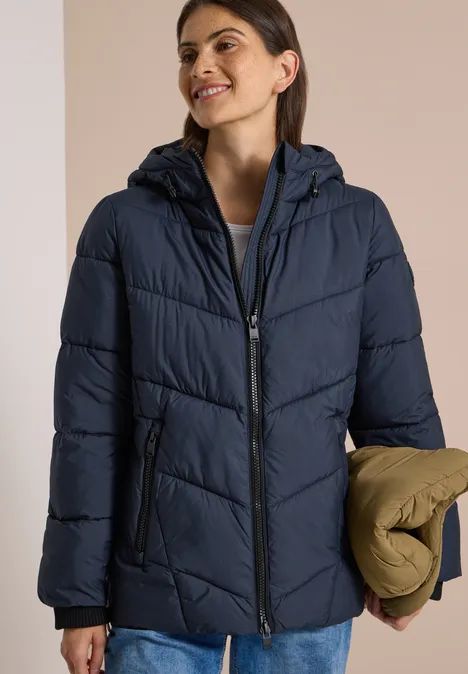 Puffer-Jacke in Midi-Länge mit Kapuze dark navy blue Frau trägt dunkelblaue Steppjacke mit Kapuze, lächelt und hält eine gefaltete khakifarbene Jacke.