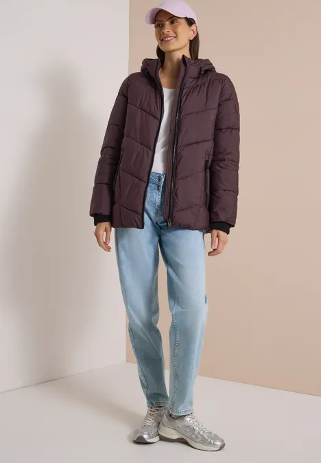 Puffer-Jacke in Midi-Länge mit Kapuze berry red Frau in lila Steppjacke, hellem Shirt, weitem Jeans, Sneakern und rosa Cap vor neutralem Hintergrund.