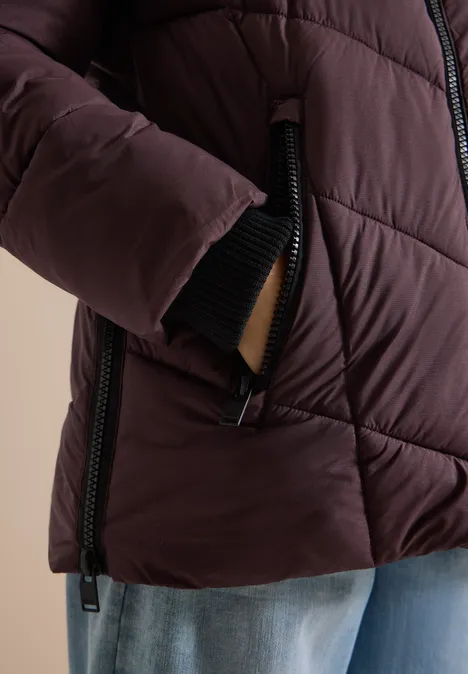 Puffer-Jacke in Midi-Länge mit Kapuze berry red Nahaufnahme einer Person mit dunkelroter Steppjacke, Hand in Tasche, schwarzer Reißverschluss, blaue Jeans.