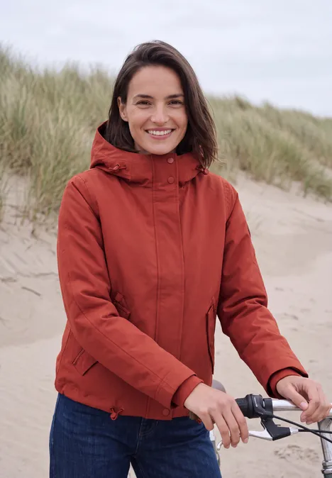 2-in-1-jas met gewatteerd vest als inzetstuk light russet red Vrouw in rode jas glimlacht op het strand en houdt fietsstuur vast, duinen op de achtergrond.