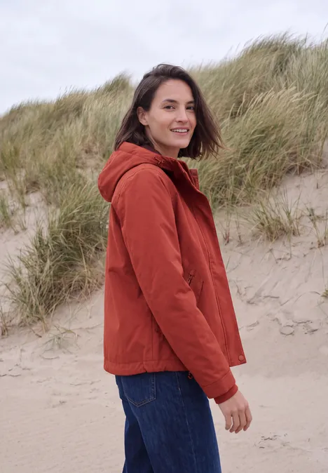 2-in-1-jas met gewatteerd vest als inzetstuk light russet red Vrouw in oranje jas op zandduin, kijkt over schouder, met helmgras op de achtergrond.