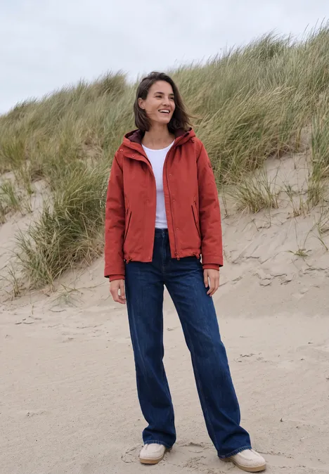 2-in-1-jas met gewatteerd vest als inzetstuk light russet red Een lachende vrouw in een rode jas, wit shirt en spijkerbroek staat voor zandduinen op het strand