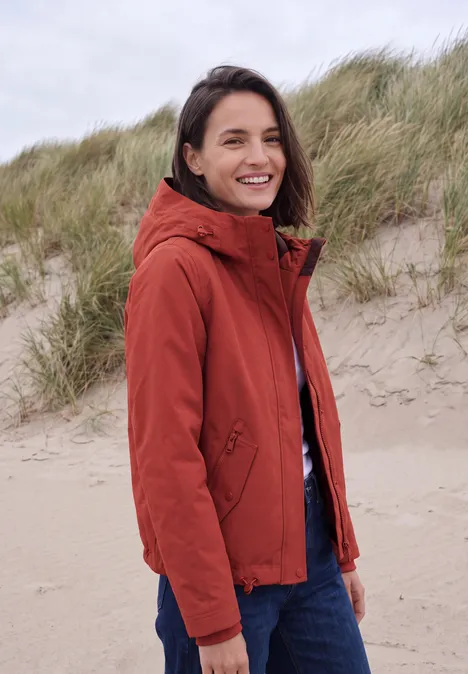 2-in-1-jas met gewatteerd vest als inzetstuk light russet red Vrouw in rood-oranje jas lacht op een winderige duinstrandlocatie.