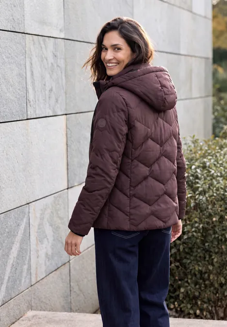 Frau in dunkelroter Steppjacke mit Kapuze steht draußen an einer Mauer und schaut lächelnd zurück.