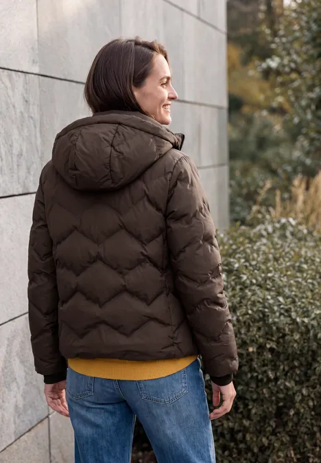 Person in brauner Daunenjacke und Jeans, lächelnd vor grauer Wand und Sträuchern.