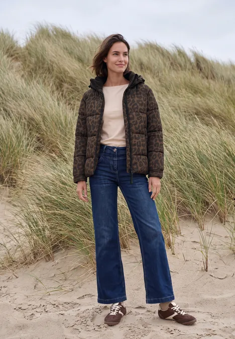 Steppjacke mit Kapuze und Reißverschluss dark toffee brown Frau in Steppjacke, Jeans und Sneakern steht auf Sand vor Dünen.
