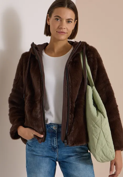 Frau in brauner Teddyjacke, weißem Shirt und Jeans trägt grüne, gesteppte Umhängetasche.