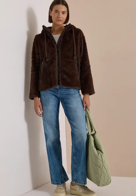 Frau in brauner, flauschiger Kapuzenjacke, blauen Jeans und mit grüner Stepp-Tasche lehnt an hellen Hintergrund.