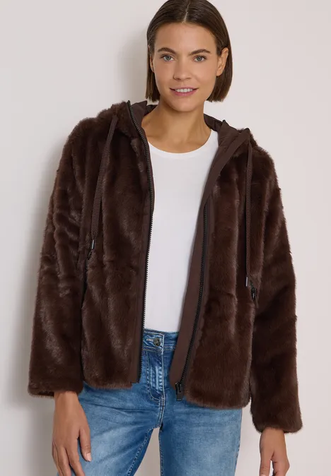 Frau in brauner, kuscheliger Kapuzenfelljacke und Jeans lächelnd.