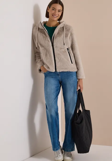 Frau lehnt lässig an der Wand mit beiger Teddyjacke, Jeans und schwarzer Tasche, lächelt freundlich.