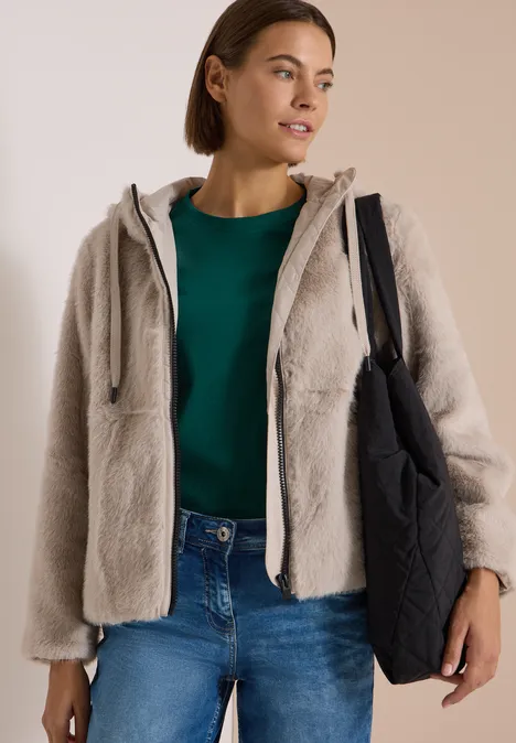 Frau in beiger Plüschjacke mit Kapuze, grünem Shirt, Jeans und großer schwarzer Tasche.
