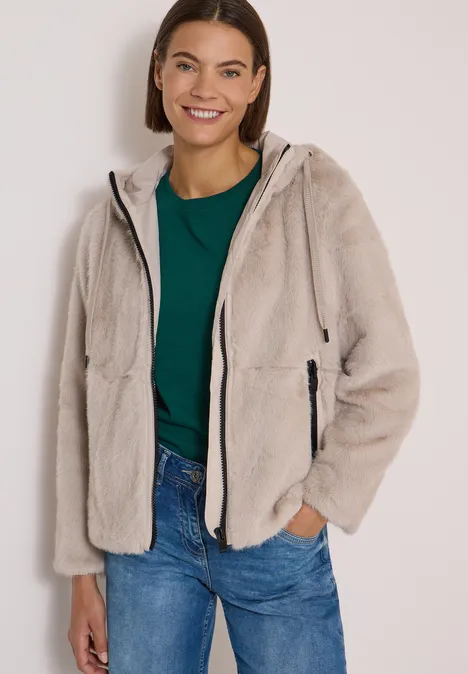 Frau mit braunen Haaren trägt eine beige Fleece-Jacke mit Kapuze, grünes Shirt und blaue Jeans.