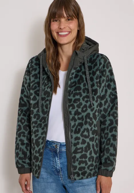 Lächelnde Frau in Jeans und weißem Shirt, trägt einen grünen Kapuzenpullover mit Leopardenmuster.