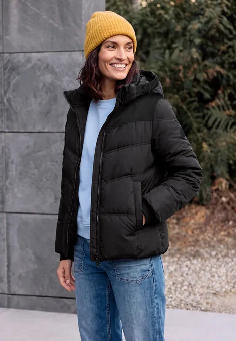 Eine lächelnde Frau trägt eine gelbe Mütze und schwarze Daunenjacke, steht lässig vor grauer Wand.