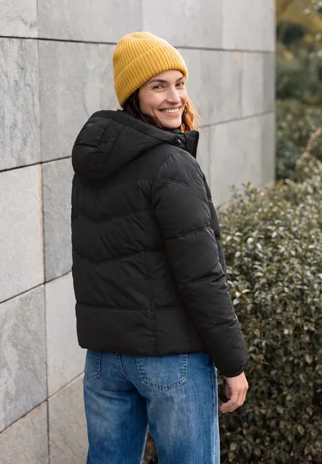 Eine Person mit gelber Strickmütze und schwarzer Steppjacke steht an einer Wand und blickt lächelnd zurück.
