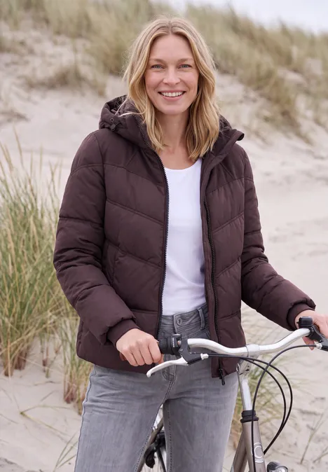 Eine lächelnde Frau in brauner Steppjacke und Jeans steht mit ihrem Fahrrad in Küstendünen.