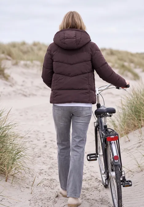 Eine Frau in einer Winterjacke schiebt ein Fahrrad über einen Dünenpfad am Strand.